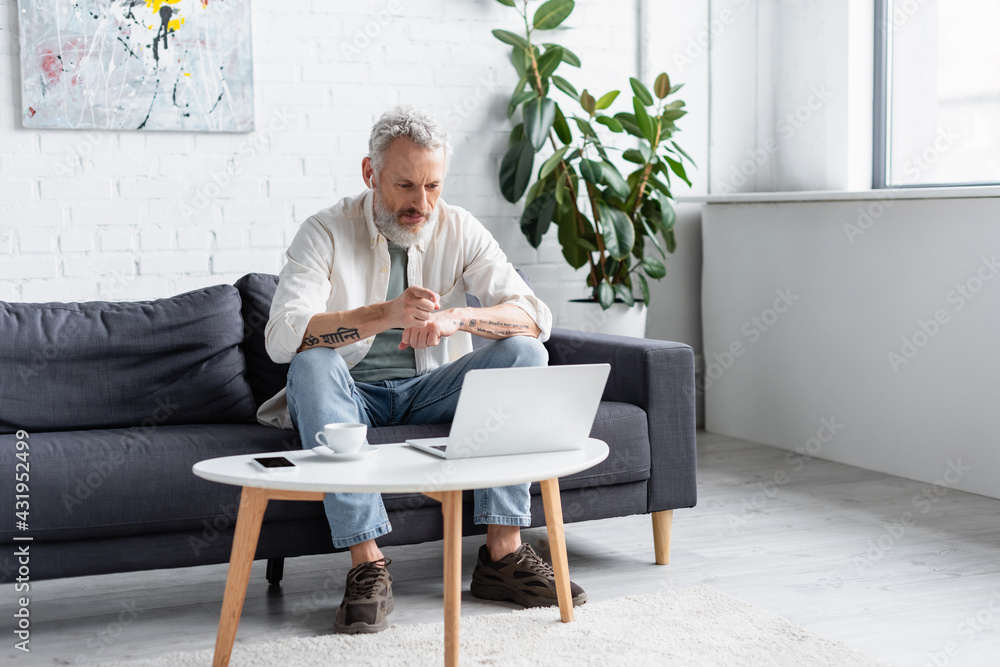 bearded man pointing at laptop while sitting on couch near cup of coffee and smartphone with blank screen on coffee table. - obrazy, fototapety, plakaty 