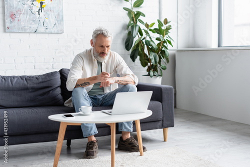 bearded man pointing at laptop while sitting on couch near cup of coffee and smartphone with blank screen on coffee table.