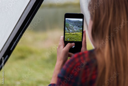 Girl taking photos of the rocky mountains and lake using the phone. Close-up shot of the handі holding the phone. View from the tent.