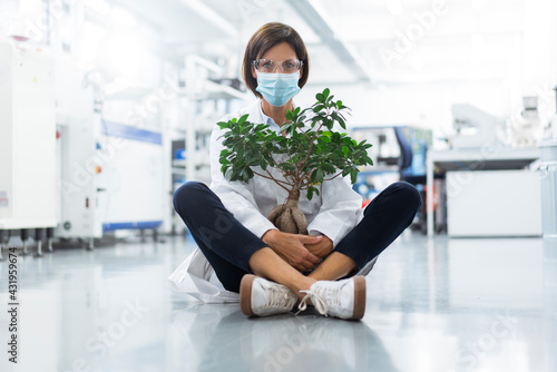 Mature female scientist with potted plant sitting in laboratory during COVID-19