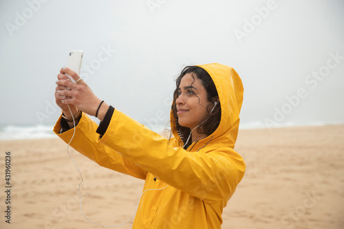 Smiling woman wearing yellow raincoat taking selfie through mobile phone while standing at beach