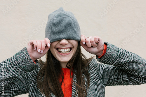 Smiling woman covering eyes through knit hat against wall photo