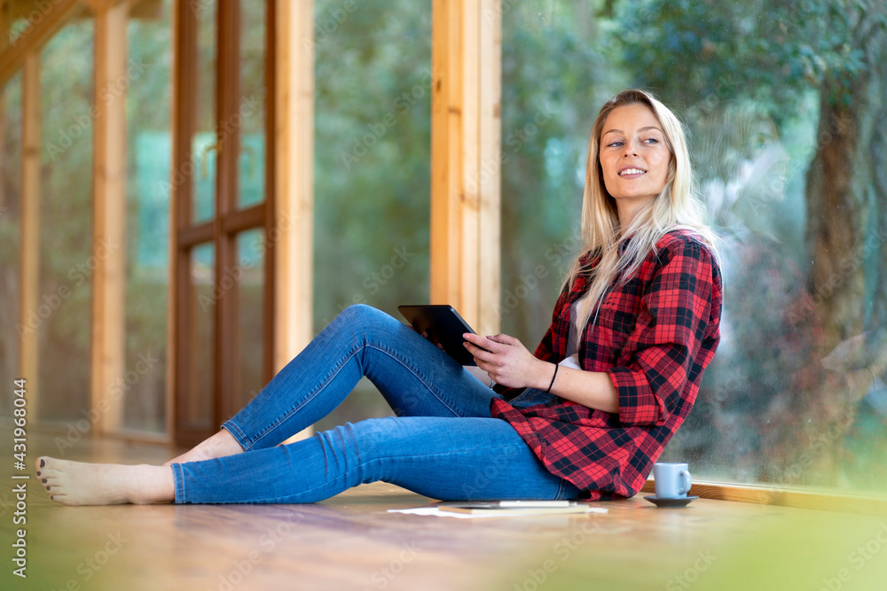 Blond woman with digital tablet looking away while sitting by window at front yard Blond woman with digital tablet looking away while sitting by window at front yard