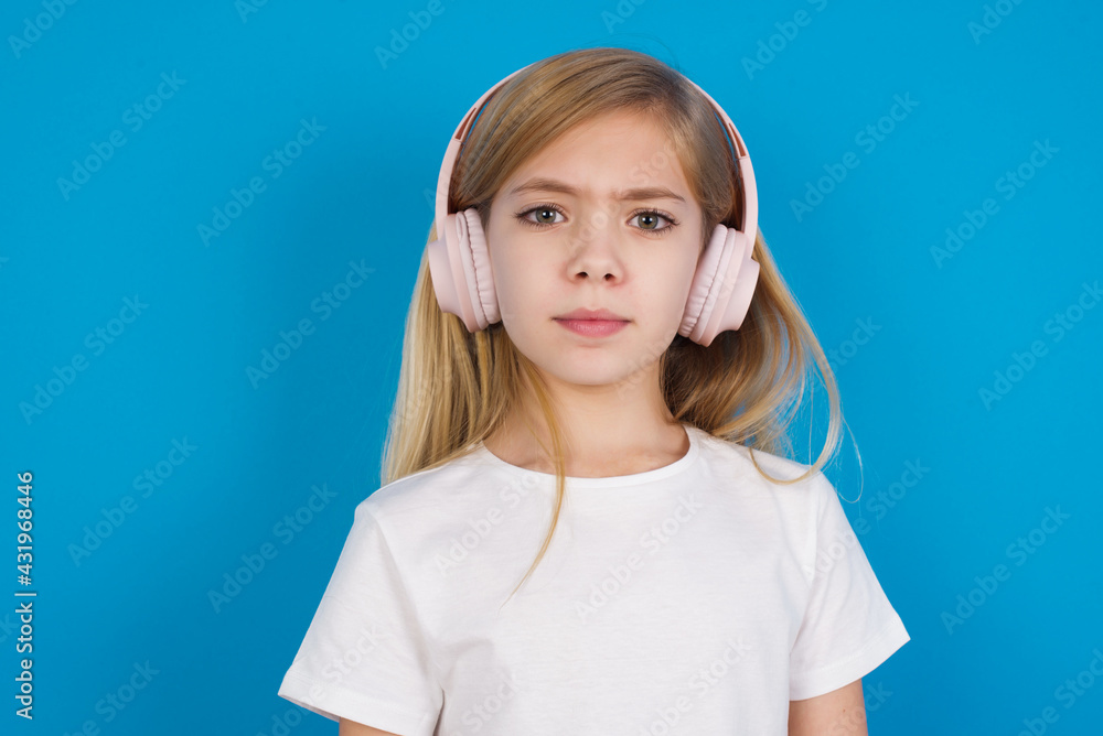 Serious displeased beautiful Caucasian little girl wearing white T-shirt over blue background looks puzzled at camera being angry wears stereo headphones listens music while walking at street Serious displeased beautiful Caucasian little girl wearing white T-shirt over blue background looks puzzled at camera being angry wears stereo headphones listens music while walking at street