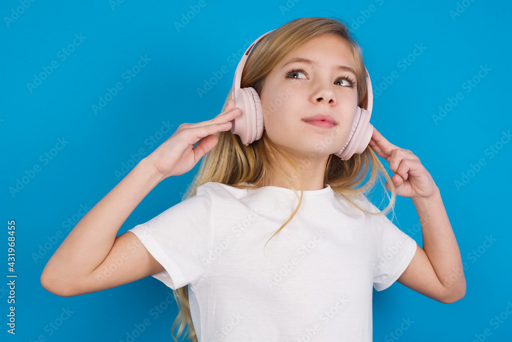 Joyful beautiful Caucasian little girl wearing white T-shirt over blue background sings song keeps hand near mouth as if microphone listens favorite playlist via headphones Joyful beautiful Caucasian little girl wearing white T-shirt over blue background sings song keeps hand near mouth as if microphone listens favorite playlist via headphones