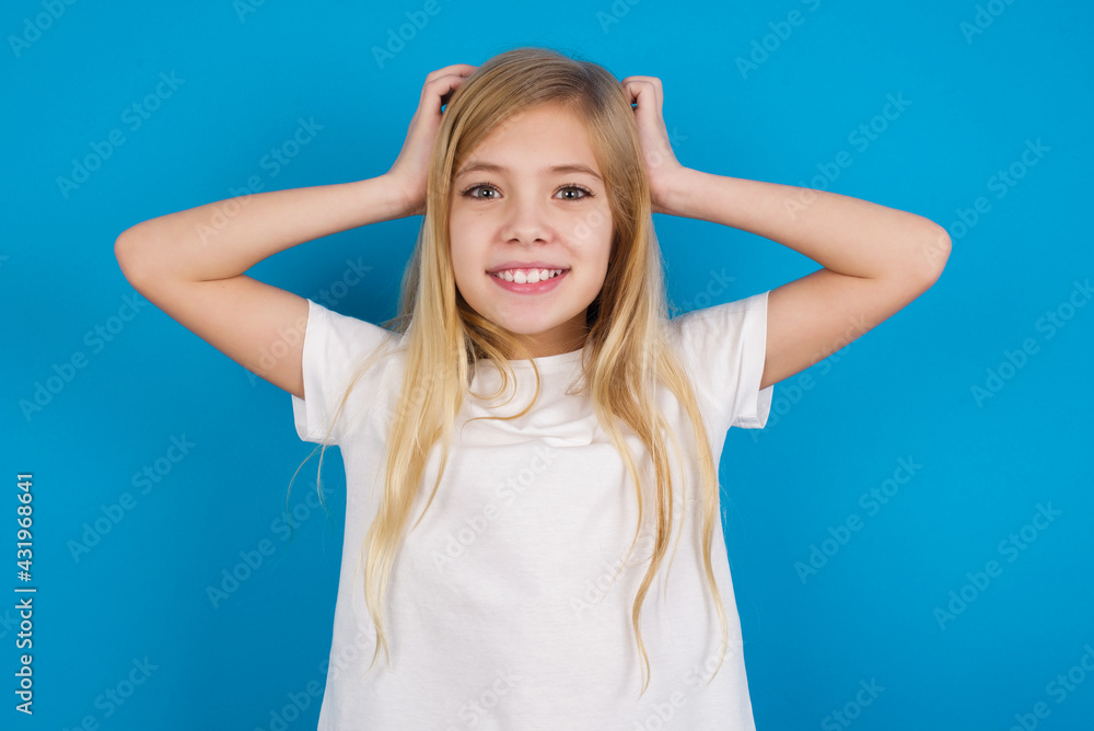 Cheerful overjoyed beautiful Caucasian little girl wearing white T-shirt over blue background reacts rising hands over head after receiving great news.