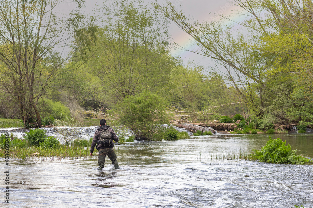 Boy fishing in the middle of a river with rocks, weeds and bushes with camouflage suit, long rubber boots, fishing rod and other tools for fishing with the rainbow in the sky