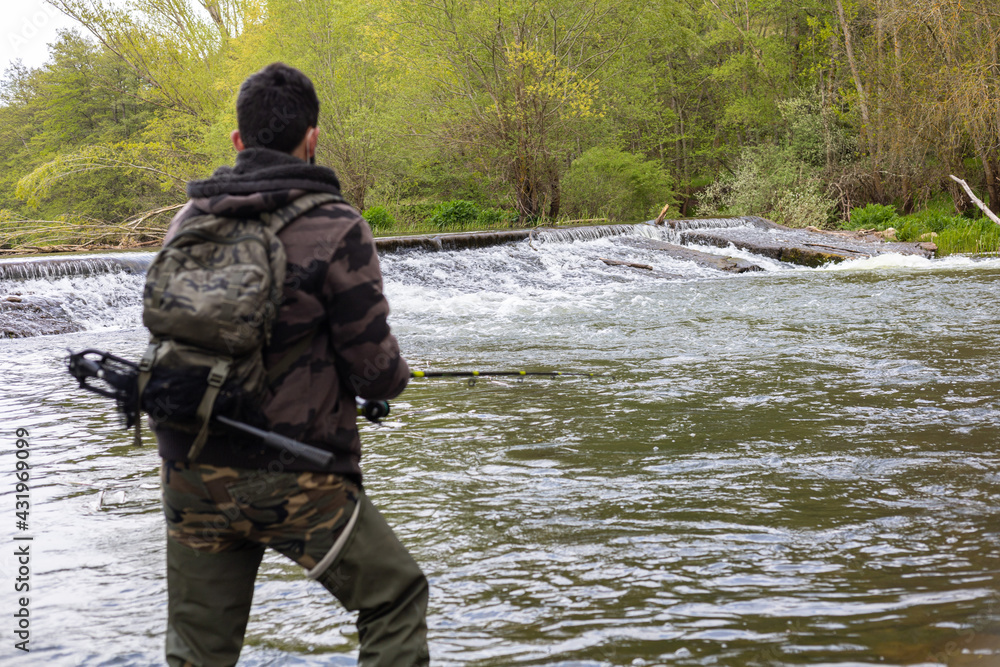 Angler with camouflage clothing, backpack with fish dryer and rubber boots fishing with a rod in a river with a small waterfall in the background
