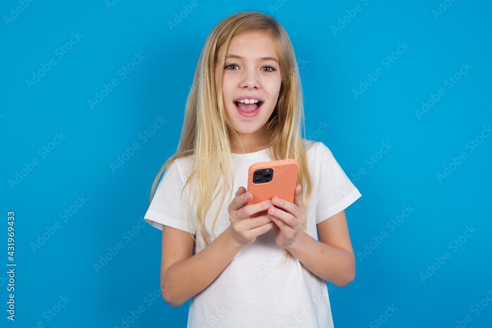 beautiful Caucasian little girl wearing white T-shirt over blue background holding gadget while sticking out tongue beautiful Caucasian little girl wearing white T-shirt over blue background holding gadget while sticking out tongue