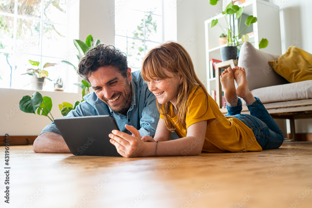 © steve brookland/Westend61 - Cheerful father and daughter looking at digital tablet while lying down on floor in living room at home