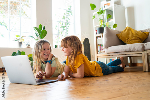 Smiling girls looking at each other while lying down on floor in front of laptop at home