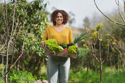 Smiling curly haired woman holding crate of leafy vegetables while standing in garden photo