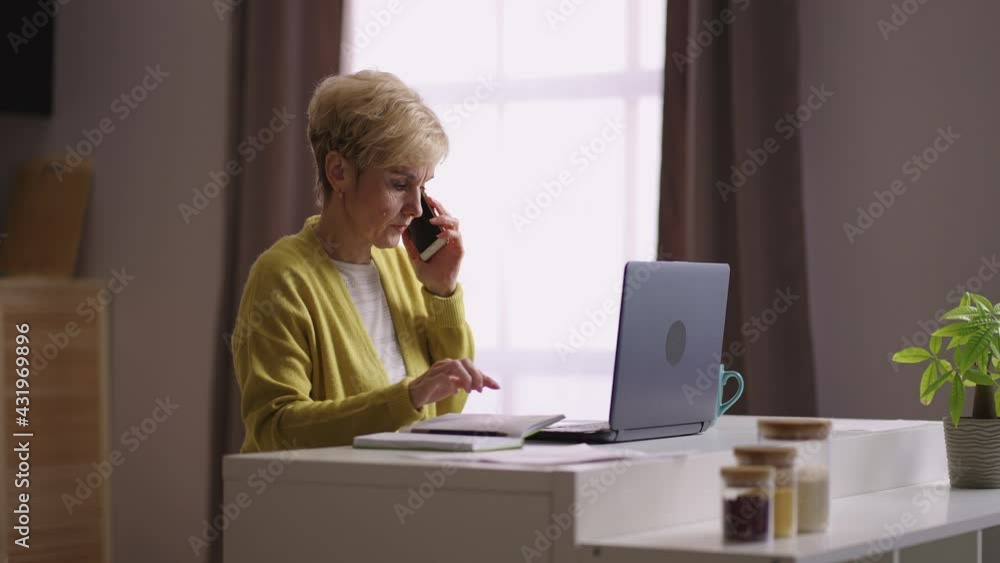old businesswoman is working in home office, communicating by phone, sitting at kitchen and using laptop old businesswoman is working in home office, communicating by phone, sitting at kitchen and using laptop
