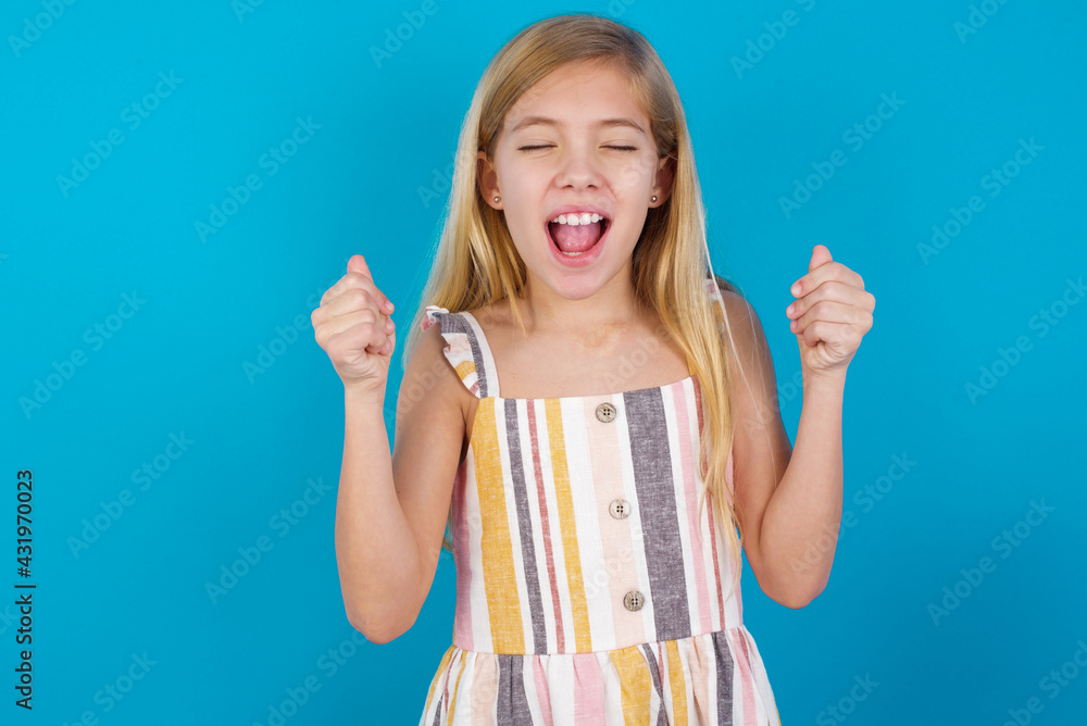 Obraz premium beautiful Caucasian little girl wearing stripped dress over blue wall rejoicing his success and victory clenching fists with joy being happy to achieve aim and goals. Positive emotions, feelings.
