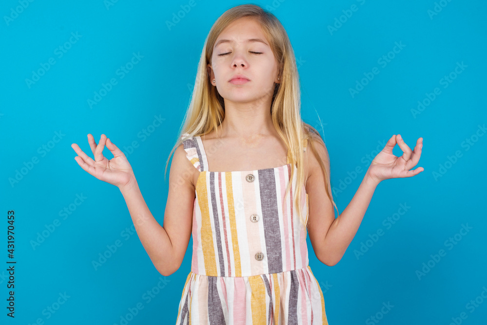beautiful Caucasian little girl wearing stripped dress over blue background doing yoga, keeping eyes closed, holding fingers in mudra gesture. Meditation, religion and spiritual practices. beautiful Caucasian little girl wearing stripped dress over blue background doing yoga, keeping eyes closed, holding fingers in mudra gesture. Meditation, religion and spiritual practices.