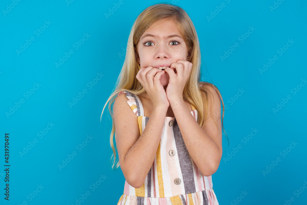 Anxiety - beautiful Caucasian little girl wearing stripped dress over blue background covering his mouth with hands scared from something or someone bitting nails. Anxiety - beautiful Caucasian little girl wearing stripped dress over blue background covering his mouth with hands scared from something or someone bitting nails.