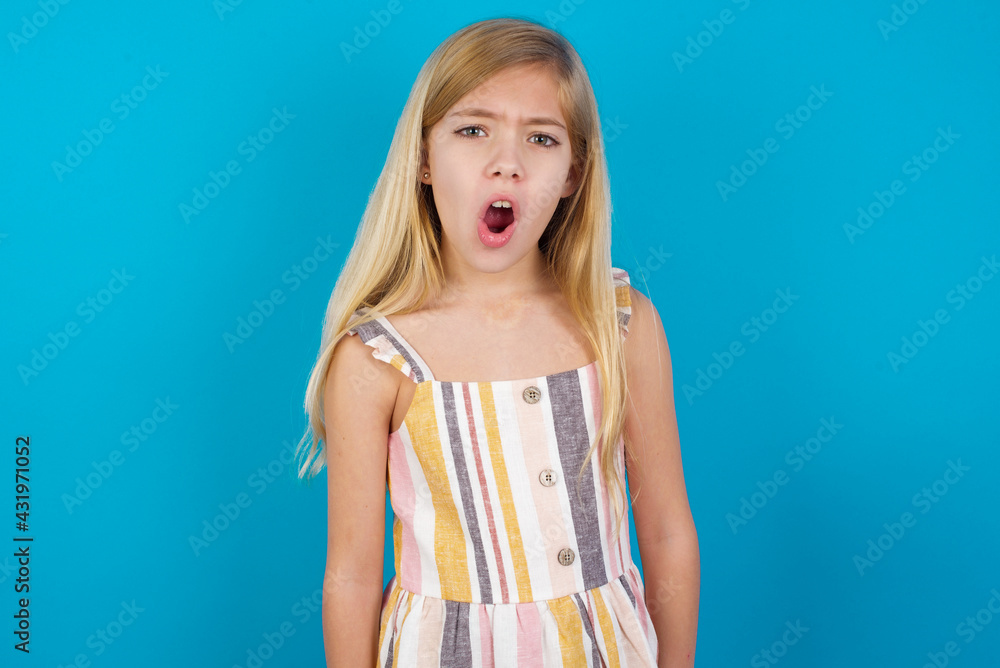 Expressive facial expressions. Shocked stupefied beautiful Caucasian little girl wearing stripped dress over blue background, keeps jaw dropped feels stunned from what he sees aside. Expressive facial expressions. Shocked stupefied beautiful Caucasian little girl wearing stripped dress over blue background, keeps jaw dropped feels stunned from what he sees aside.