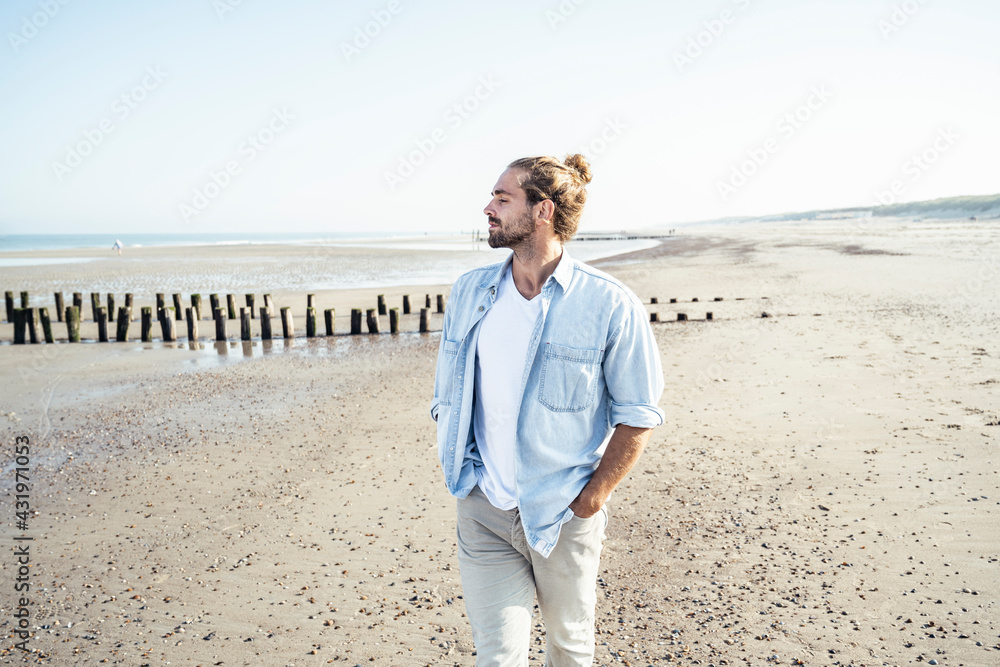 Handsome man with hands in pockets standing at beach on sunny day Handsome man with hands in pockets standing at beach on sunny day