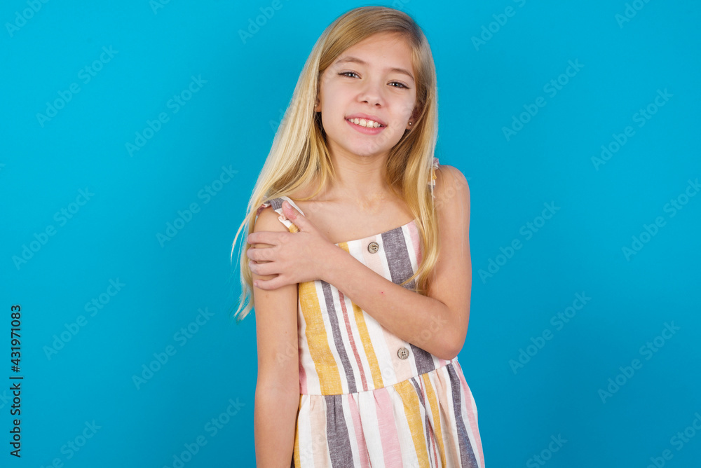 People, lifestyle, youth and happiness concept. Shy pretty beautiful Caucasian little girl wearing stripped dress over blue background, feeling happy hugging herself. People, lifestyle, youth and happiness concept. Shy pretty beautiful Caucasian little girl wearing stripped dress over blue background, feeling happy hugging herself.