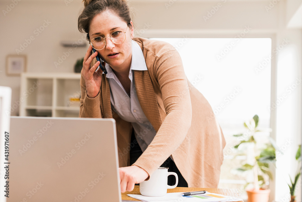 Female entrepreneur talking on smart phone while looking at laptop Female entrepreneur talking on smart phone while looking at laptop