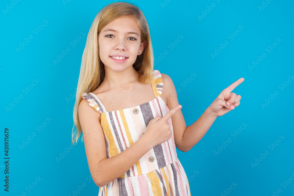 beautiful Caucasian little girl wearing stripped dress over blue background with positive expression, indicates with fore finger at blank copy space for your promotional text or advertisement. beautiful Caucasian little girl wearing stripped dress over blue background with positive expression, indicates with fore finger at blank copy space for your promotional text or advertisement.