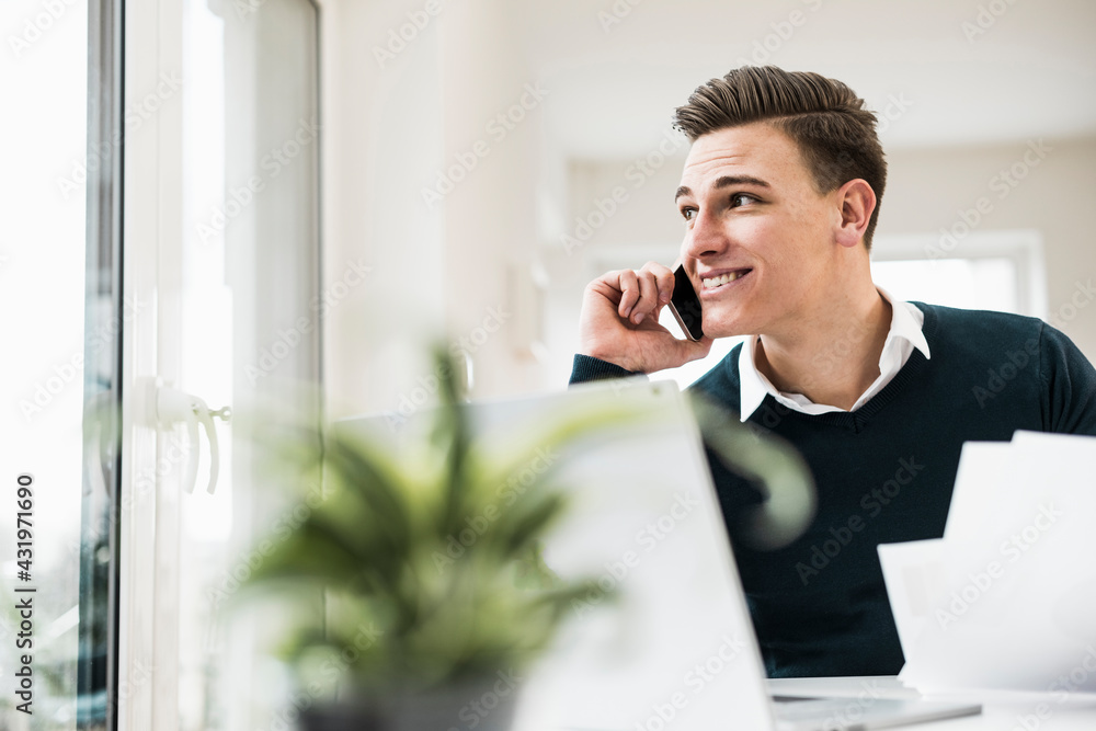 Smiling male entrepreneur talking on phone while looking through window at home office Smiling male entrepreneur talking on phone while looking through window at home office