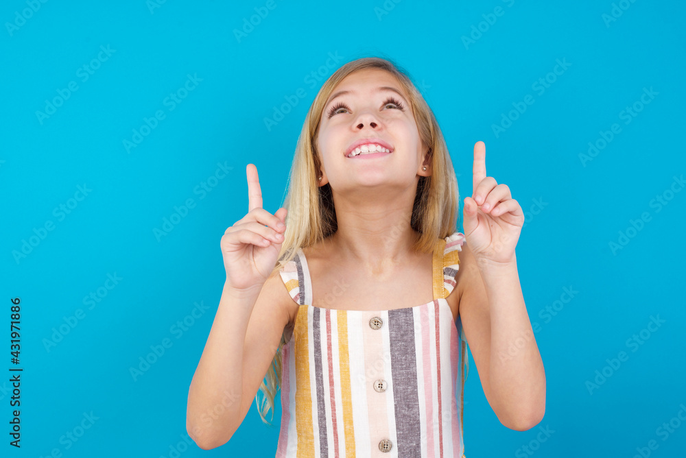 Successful friendly looking beautiful Caucasian little girl wearing stripped dress over blue background exclaiming excitedly, pointing both index fingers up, indicating something. Successful friendly looking beautiful Caucasian little girl wearing stripped dress over blue background exclaiming excitedly, pointing both index fingers up, indicating something.