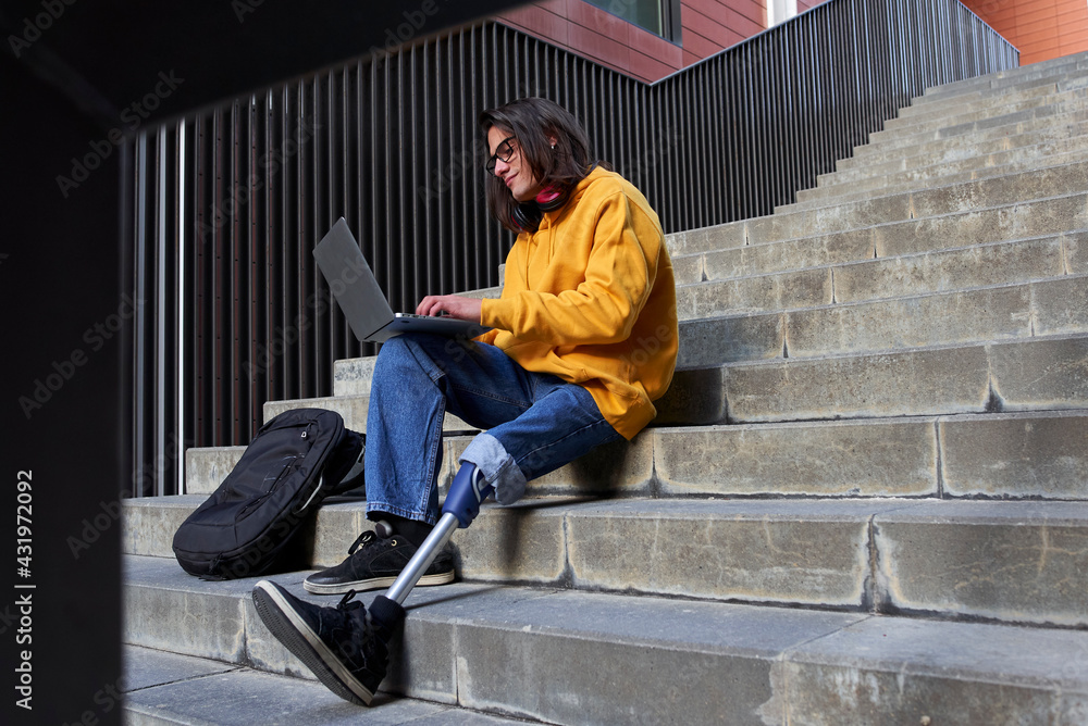 Disabled young man with leg prosthesis using laptop while sitting on steps Disabled young man with leg prosthesis using laptop while sitting on steps