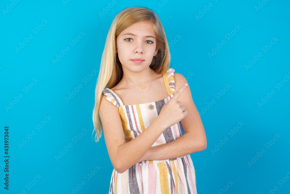 beautiful Caucasian little girl wearing stripped dress over blue background smiling broadly at camera, pointing fingers away, showing something interesting and exciting. beautiful Caucasian little girl wearing stripped dress over blue background smiling broadly at camera, pointing fingers away, showing something interesting and exciting.