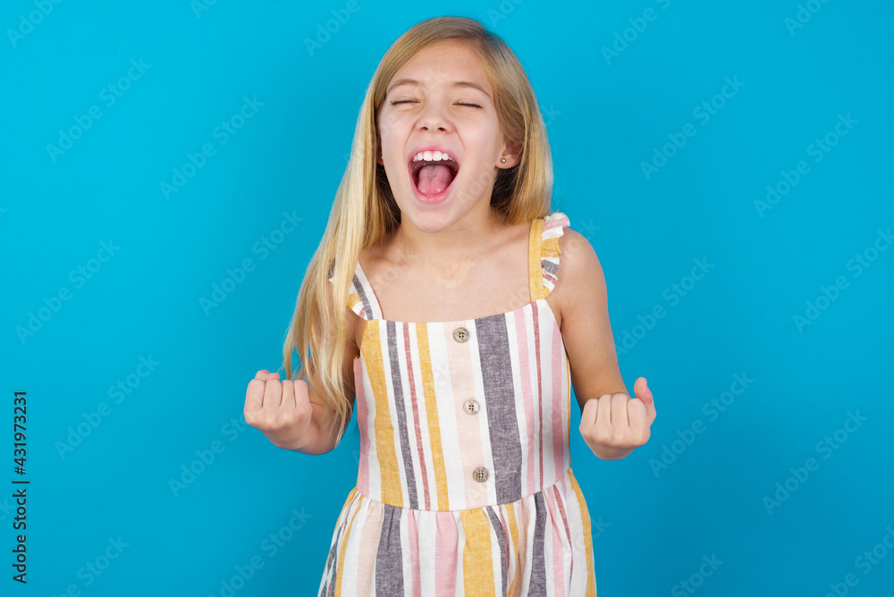Portrait of beautiful Caucasian little girl wearing stripped dress over blue looks with excitement at camera, keeps hands raised over head, notices something unexpected reacts on sudden news. Portrait of beautiful Caucasian little girl wearing stripped dress over blue looks with excitement at camera, keeps hands raised over head, notices something unexpected reacts on sudden news.
