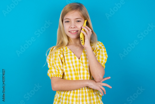 Portrait of a smiling beautiful Caucasian little girl wearing yellow dress over blue background talking on mobile phone. Business, confidence and communication concept. © Jihan