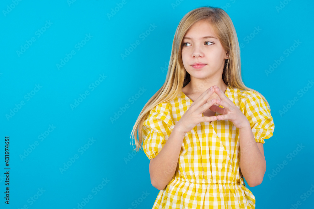 beautiful Caucasian little girl wearing yellow dress over blue background steepls fingers and looks mysterious aside has great evil plan in mind beautiful Caucasian little girl wearing yellow dress over blue background steepls fingers and looks mysterious aside has great evil plan in mind