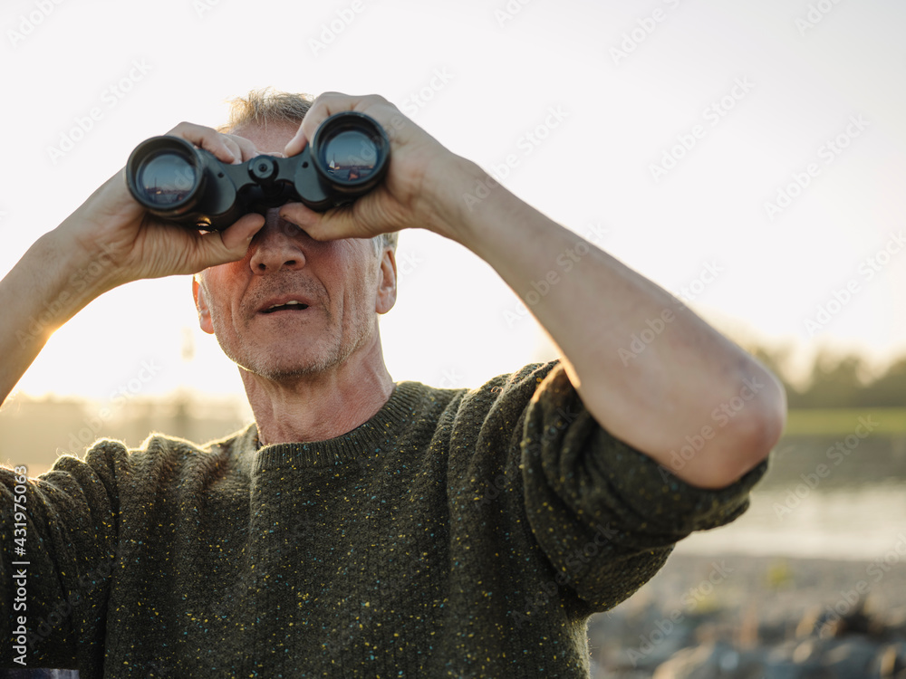 Senior man looking through binoculars against clear sky at riverbank Senior man looking through binoculars against clear sky at riverbank