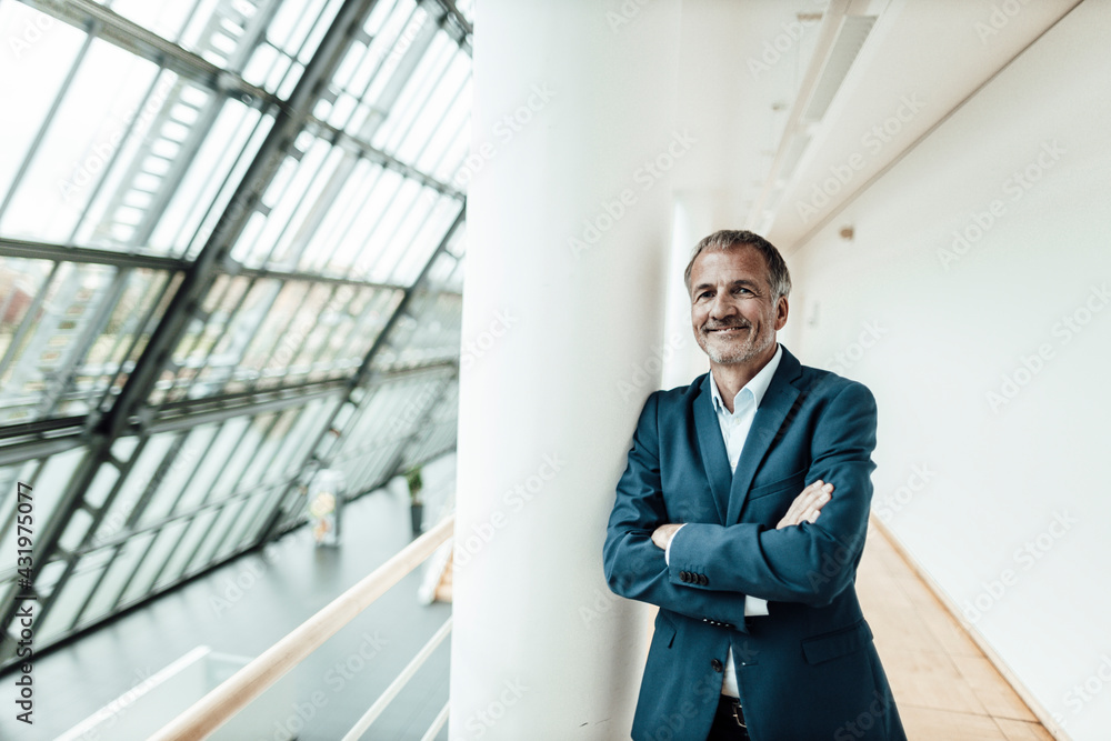 Smiling businessman with arms crossed standing at column in office corridor Smiling businessman with arms crossed standing at column in office corridor