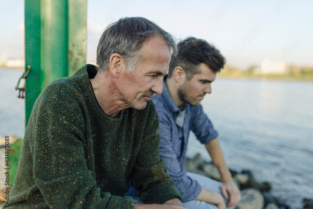 © Gustafsson/Westend61 - Son and father looking down while sitting by river