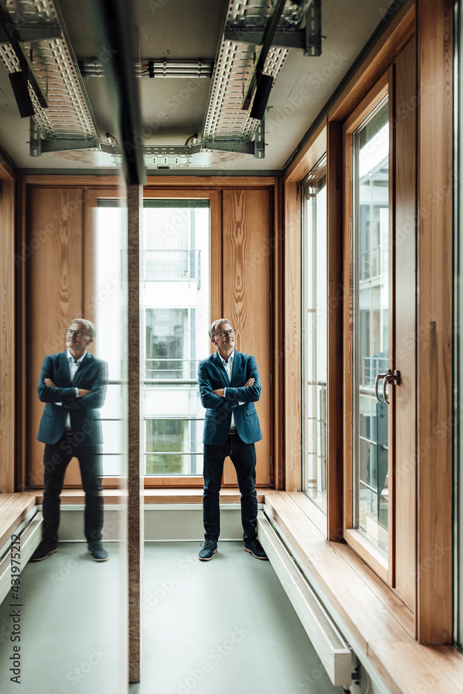 Male entrepreneur with arms crossed standing in office corridor Male entrepreneur with arms crossed standing in office corridor