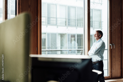 Smiling senior male entrepreneur with arms crossed looking through glass window in office