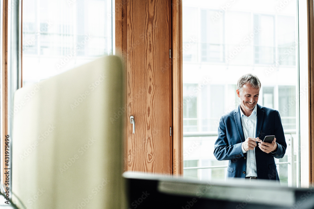 Smiling senior businessman using smart phone while standing against glass window in office Smiling senior businessman using smart phone while standing against glass window in office