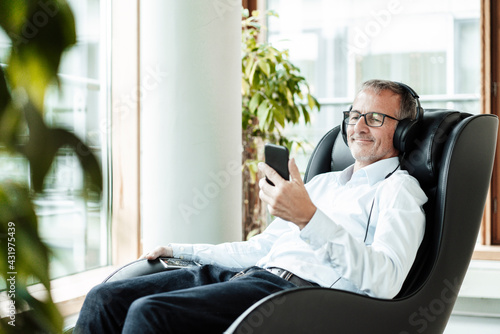 Smiling male business professional using smart phone while listening music through headphones on massage chair photo
