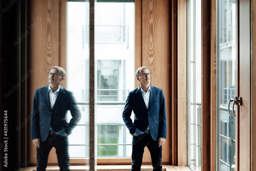 Male business professional with hand in pocket contemplating while standing against glass window at office corridor Male business professional with hand in pocket contemplating while standing against glass window at office corridor