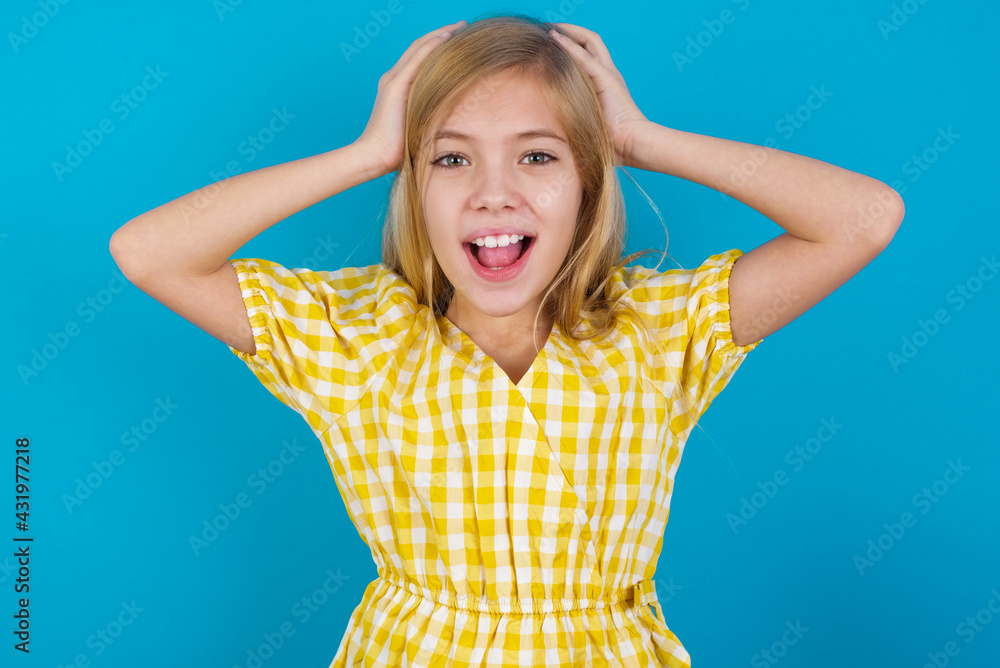 Cheerful overjoyed beautiful Caucasian little girl wearing yellow dress over blue background reacts rising hands over head after receiving great news.