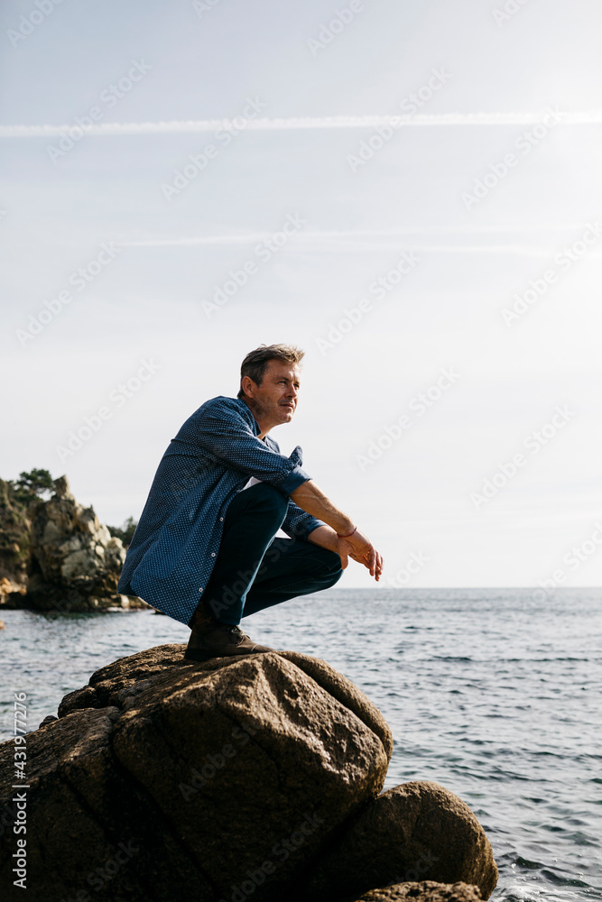 Mature man crouching on rock by sea against clear sky
