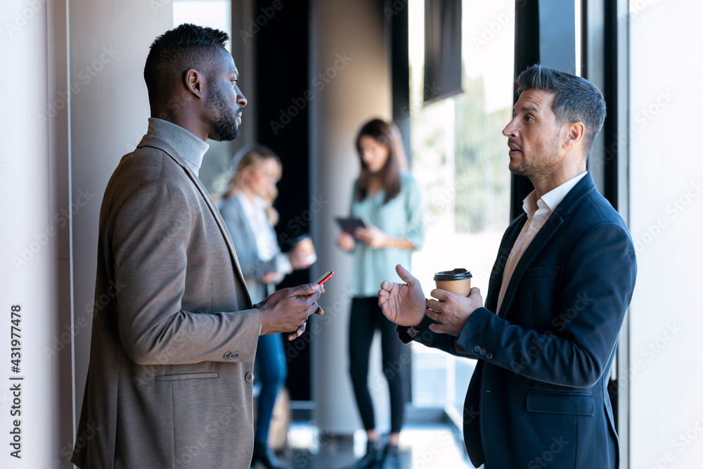 Businessmen having discussion while standing with colleague in background at office Businessmen having discussion while standing with colleague in background at office