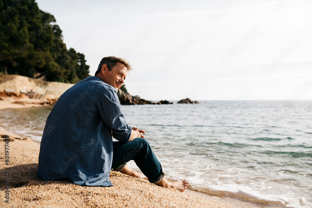 Smiling man sitting on beach while looking away against sea at beach Smiling man sitting on beach while looking away against sea at beach