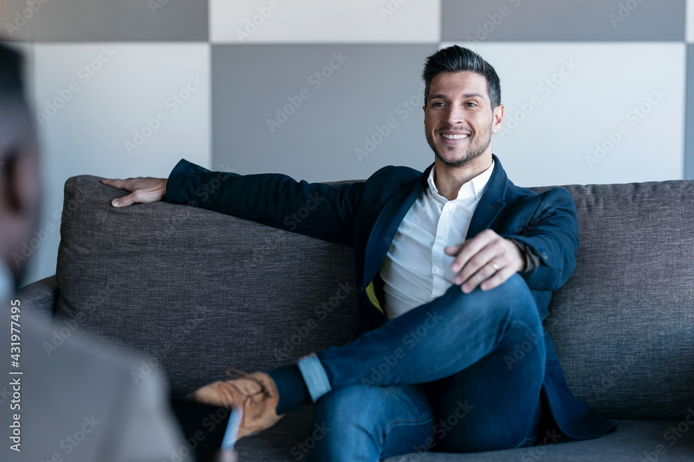 Confident businessman talking to colleague while sitting with legs crossed at knee on couch at office Confident businessman talking to colleague while sitting with legs crossed at knee on couch at office