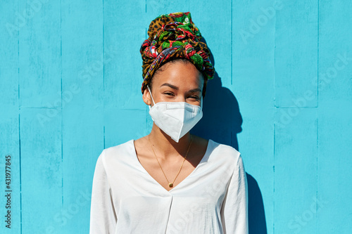 Woman wearing protective face mask and headscarf staring while standing against blue wall photo