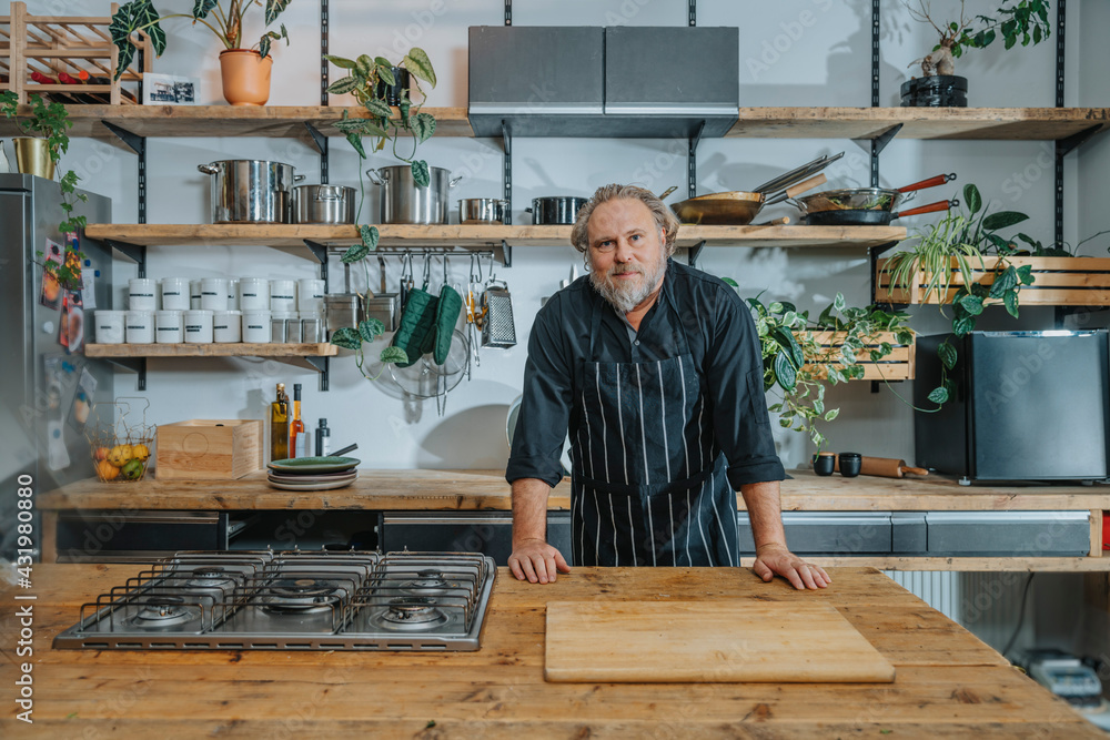 Confident mature chef staring while standing by kitchen counter in kitchen Confident mature chef staring while standing by kitchen counter in kitchen