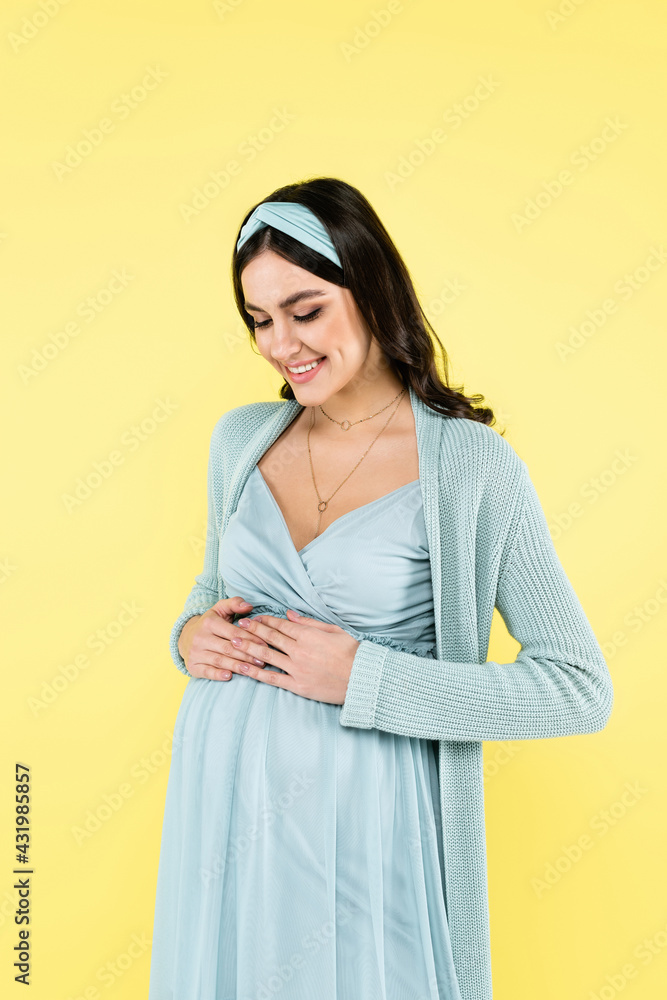 joyful pregnant woman holding hands on tummy while smiling isolated on yellow. joyful pregnant woman holding hands on tummy while smiling isolated on yellow.