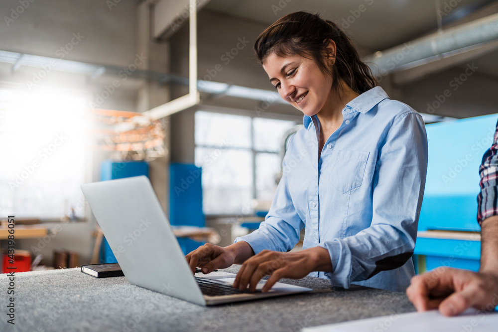 Smiling businesswoman working on laptop while standing by colleague at industry Smiling businesswoman working on laptop while standing by colleague at industry