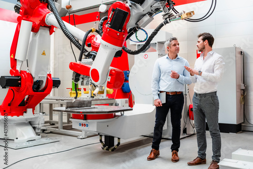 Male engineers discussing about plans by robotics in factory photo